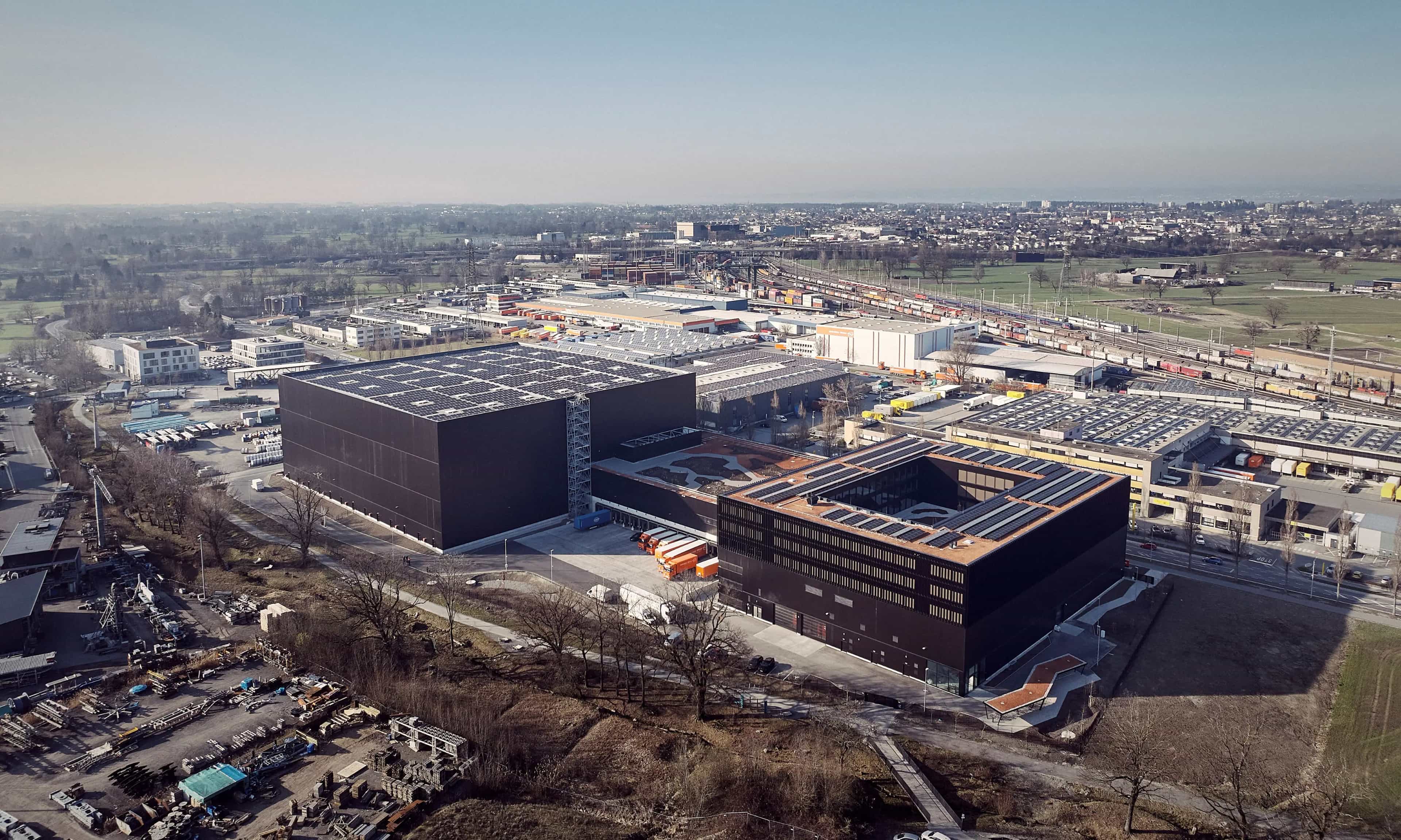Luftaufnahme eines großen Industriekomplexes mit schwarzen Gebäuden und umliegendem Gelände in einer städtischen Umgebung.