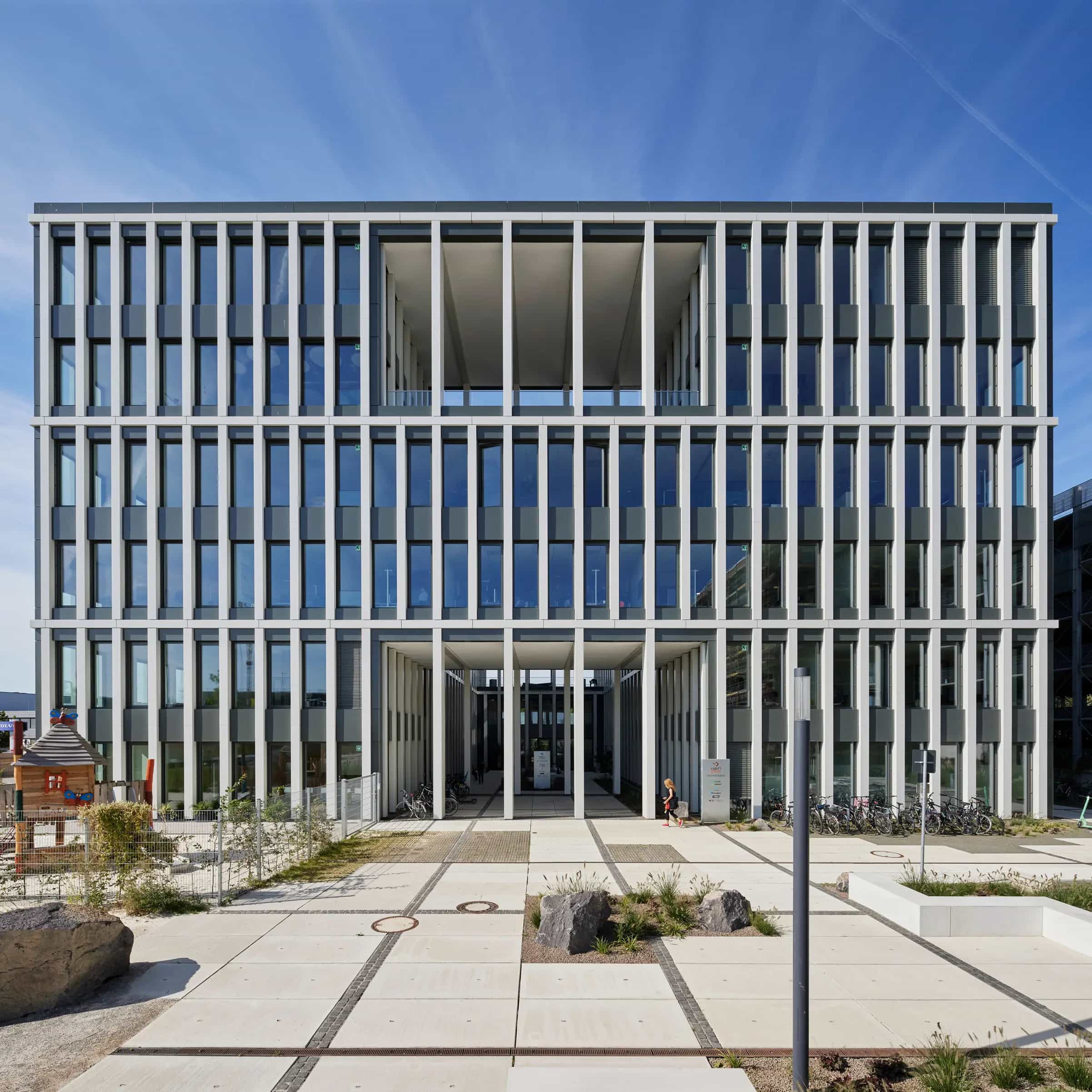 Moderne Fassade eines Bürogebäudes mit großen Fenstern und klaren Linien. Im Vordergrund eine gepflasterte Fläche und ein Spielbereich.