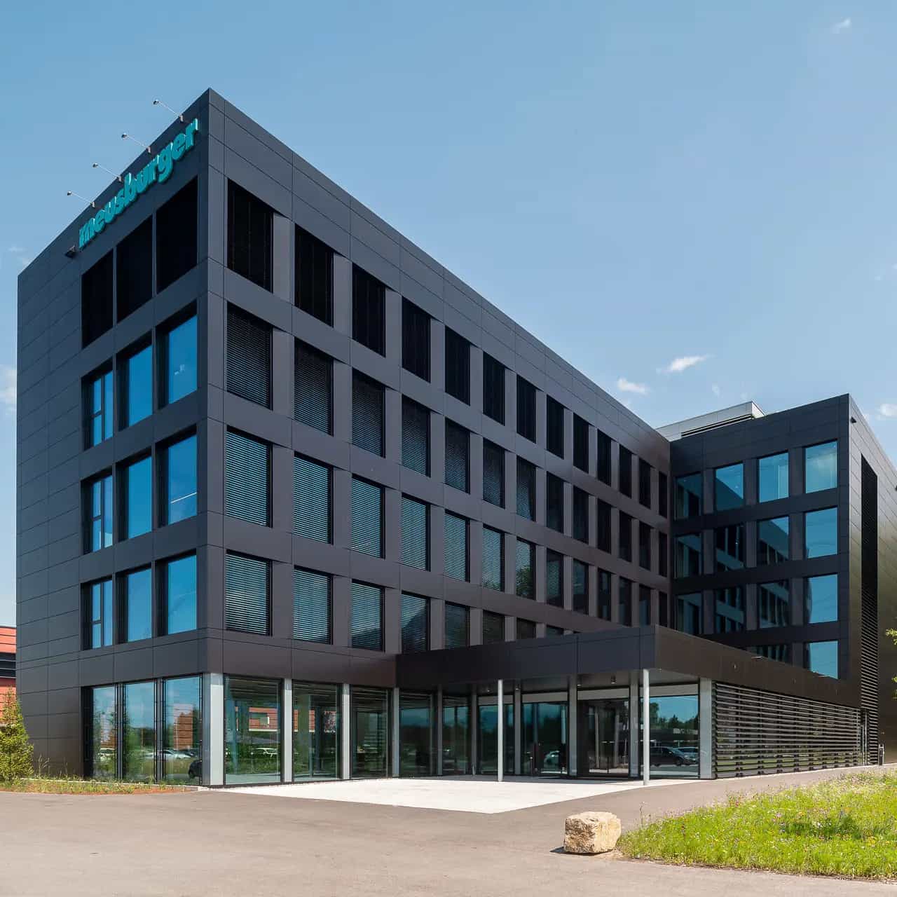 Modern office building with glass façade under a blue sky, surrounded by green spaces.