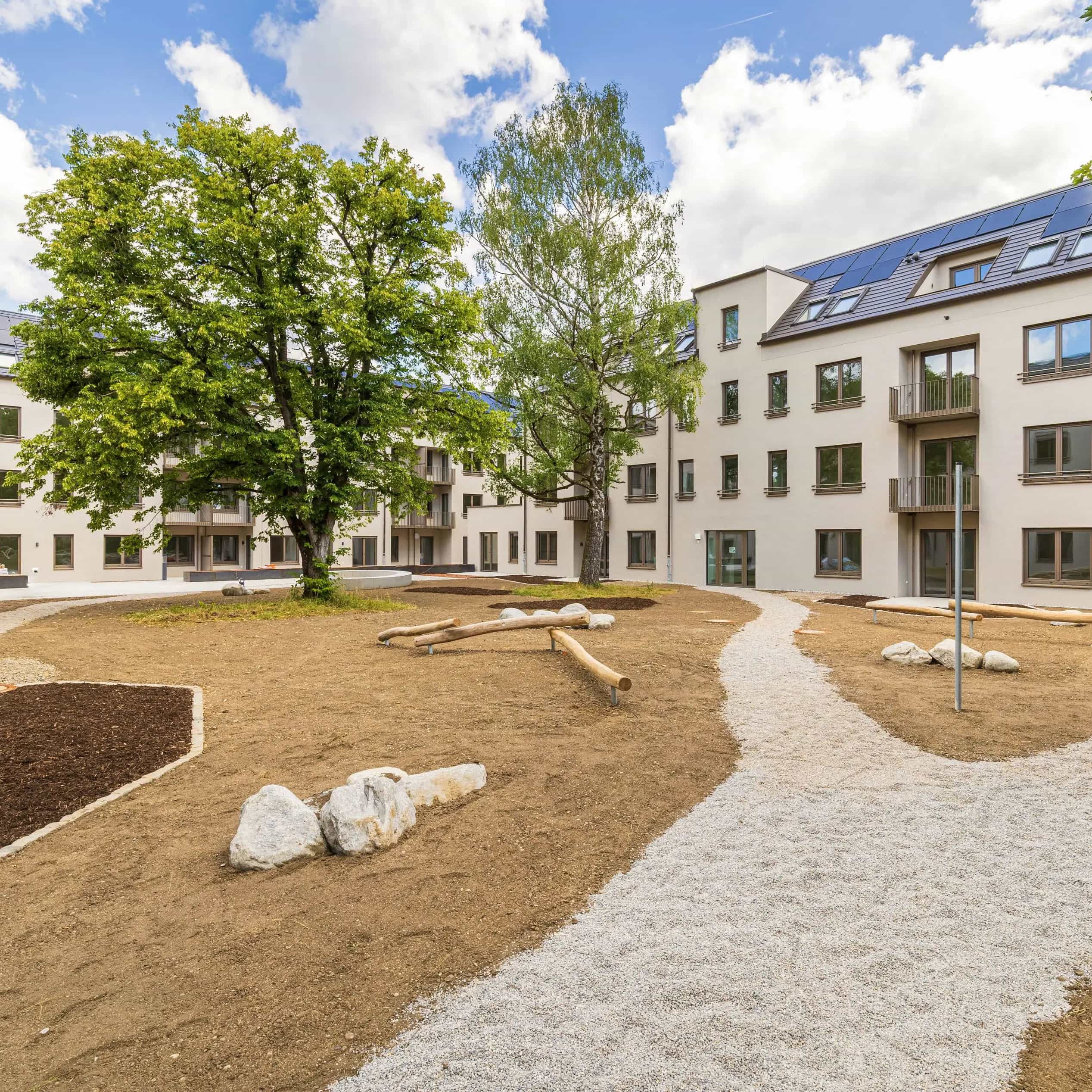 A new residential complex with a green courtyard, gravel paths, and trees under a blue sky.