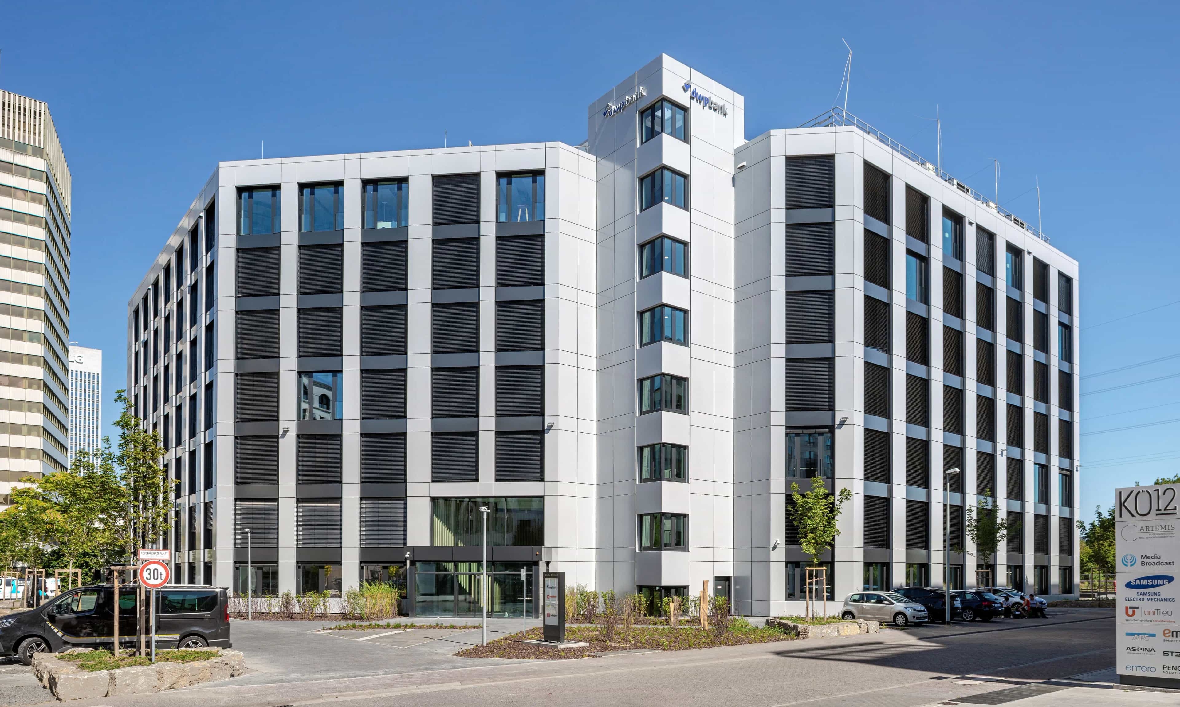 A modern office building with a glass facade under a clear blue sky, surrounded by trees and parked cars.