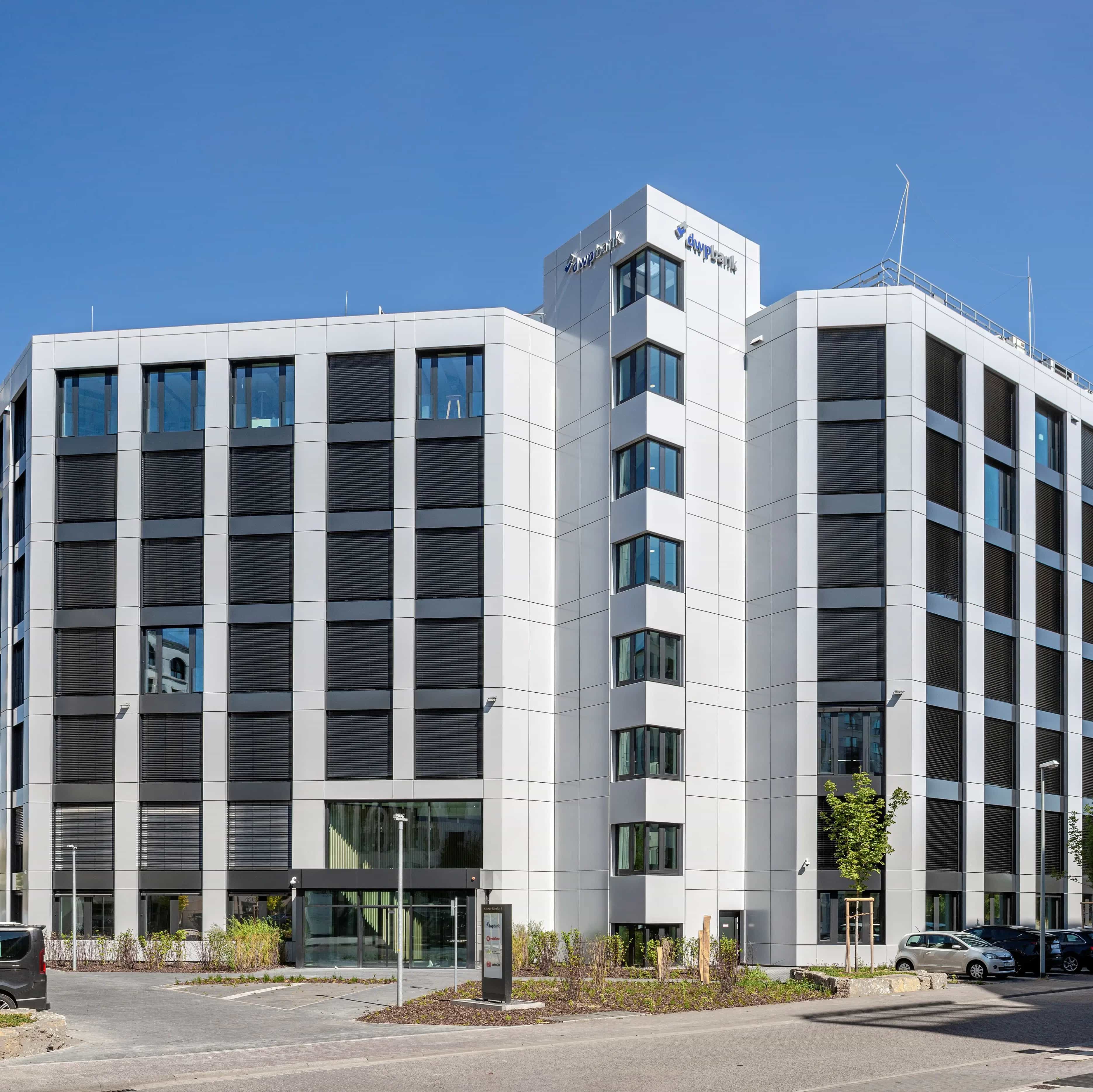 A modern office building with a glass facade under a clear blue sky, surrounded by trees and parked cars.